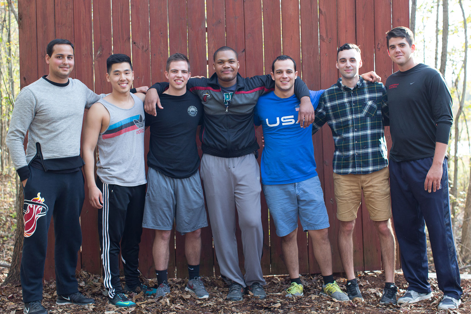 Group of men in casual attire next to a tall fence in the woods.