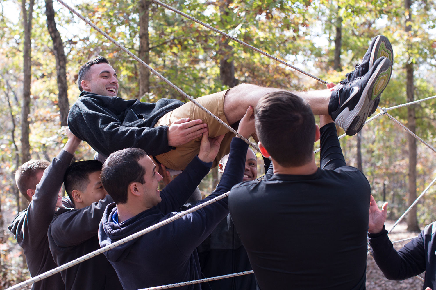 Group of young men holiding up one man and passing him through a web of ropes in a woods setting.