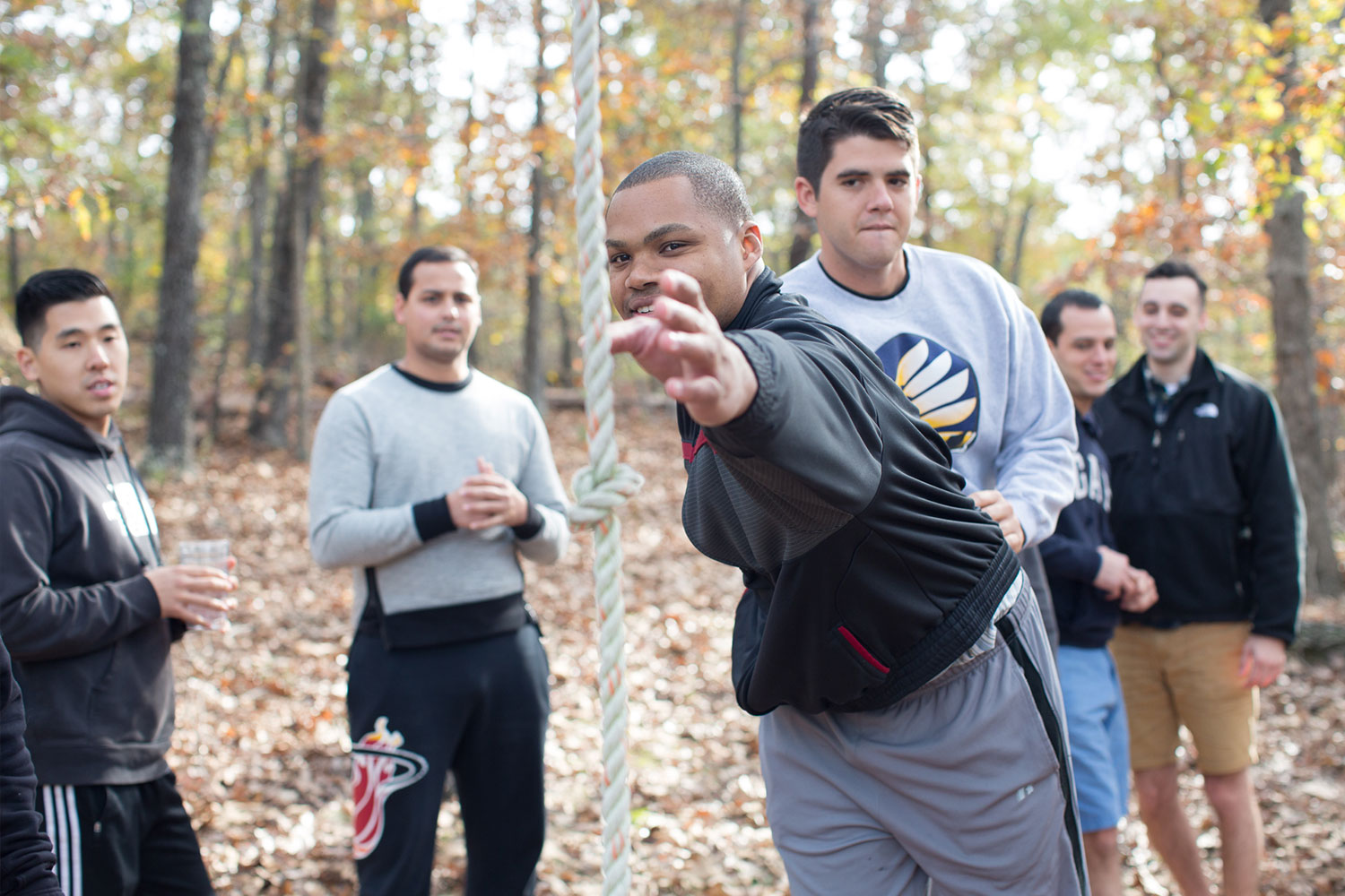 Group of young men in the woods and one man reaching for a rope knot in the foreground.