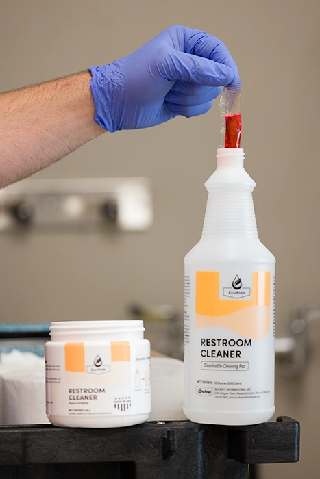 A gloved hand dropping an Eco Pod into Restroom Cleaner Spray Bottle